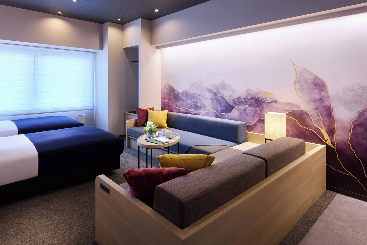 Habitación de hotel con dos camas, un sofá, una mesita con lámpara y un adorno en la pared.