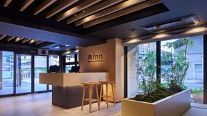 Um balcão de recepção moderno com uma placa indicando 'Minn sua segunda casa', dois bancos de madeira, uma janela grande com vista para edifícios e vegetação, e um teto com ripas de madeira.