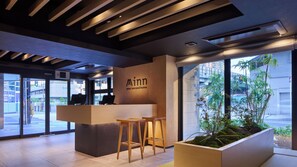 A modern reception desk with a sign reading 'Minn your second home,' two wooden stools, a large window with a view of buildings and greenery, and a ceiling with wooden slats. - Minn Namba South (Osaka)