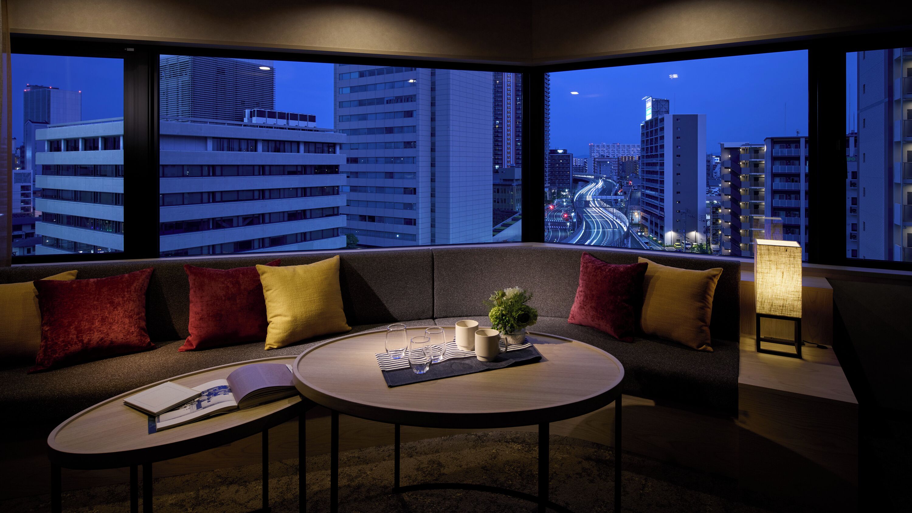 A modern hotel room with a large window, a sofa, a small round table, and a lamp.