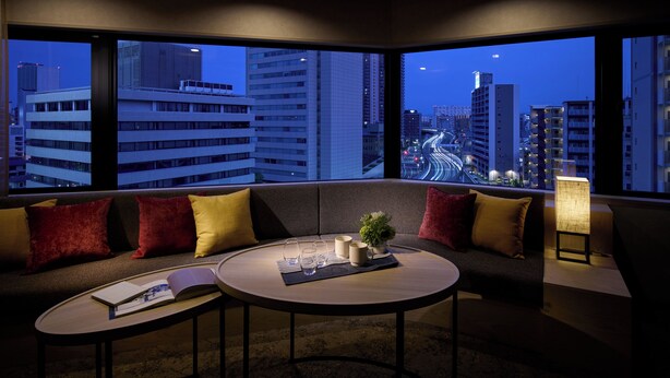 Quarto de hotel moderno com uma grande janela, um sofá, uma pequena mesa redonda e um abajur.