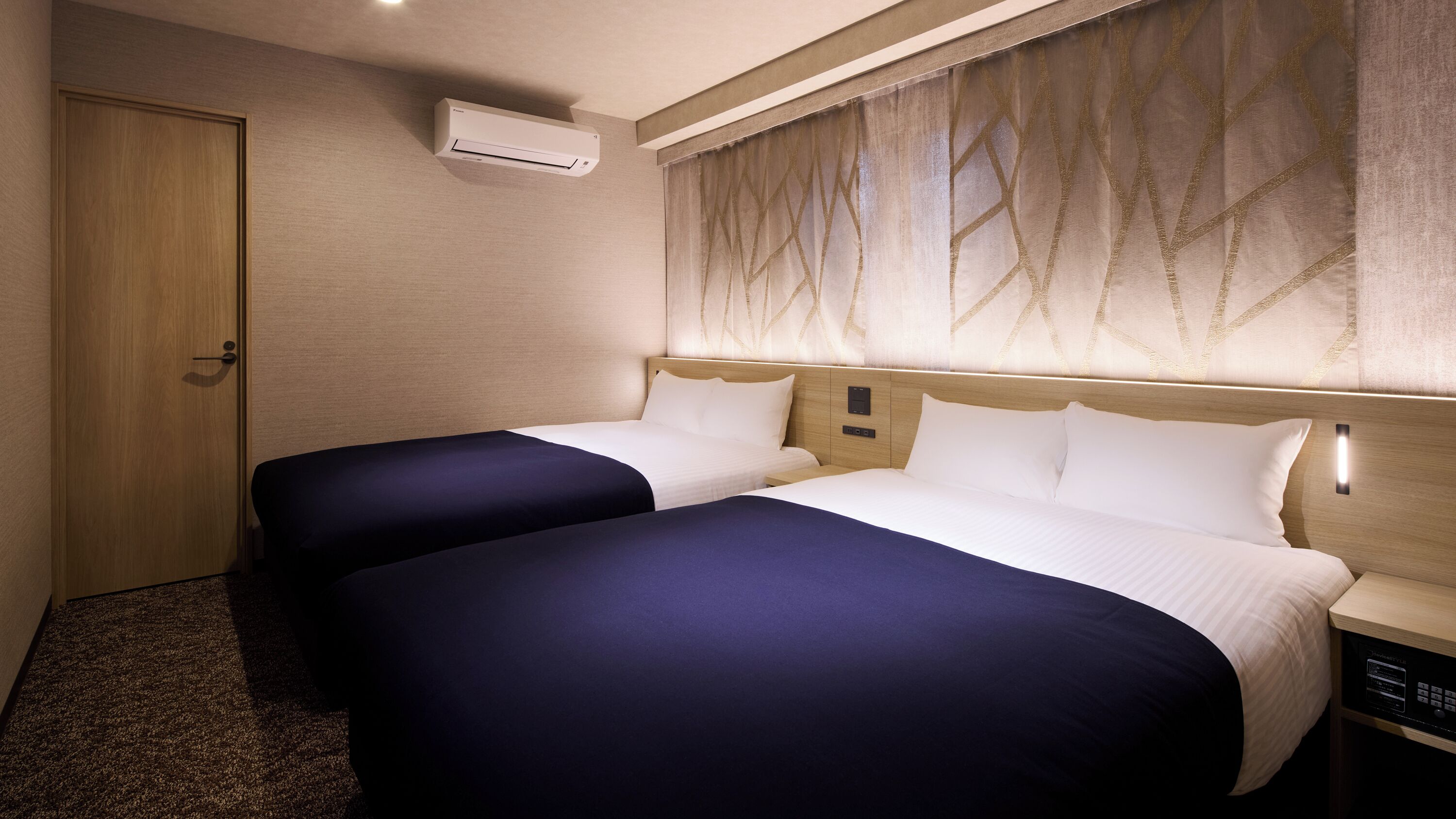 Habitación de hotel con dos camas, un equipo de aire acondicionado, una puerta de madera y una cortina con estampado de hojas.