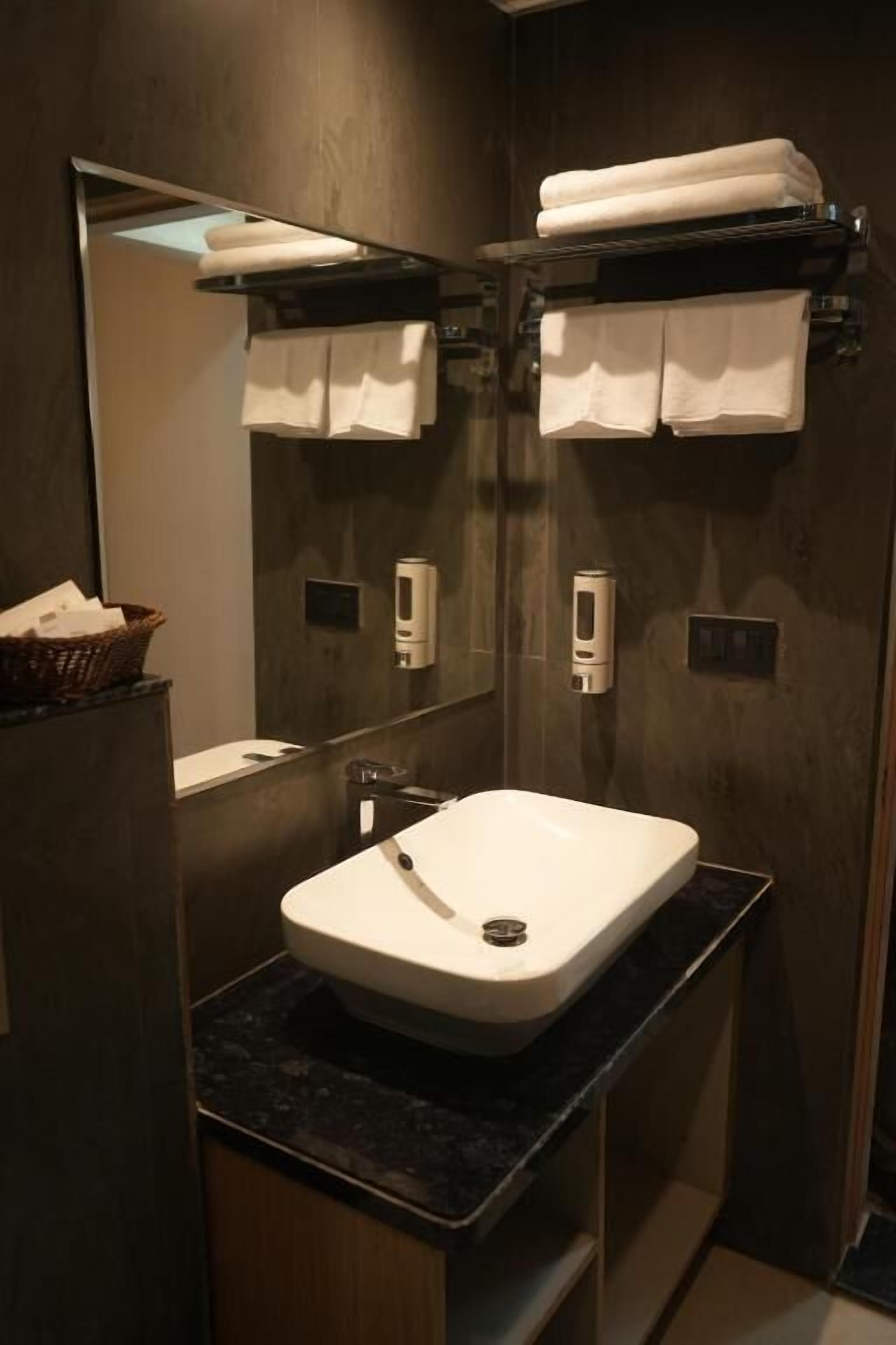 A modern bathroom with a white sink, dark countertop, and a mirror above. There are two towels on a shelf and a basket on the left.