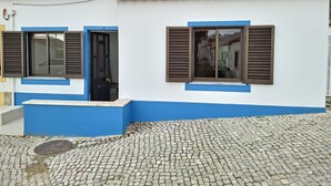 Un edificio con tetto in tegole, due finestre con persiane, un ingresso e un terreno lastricato.
