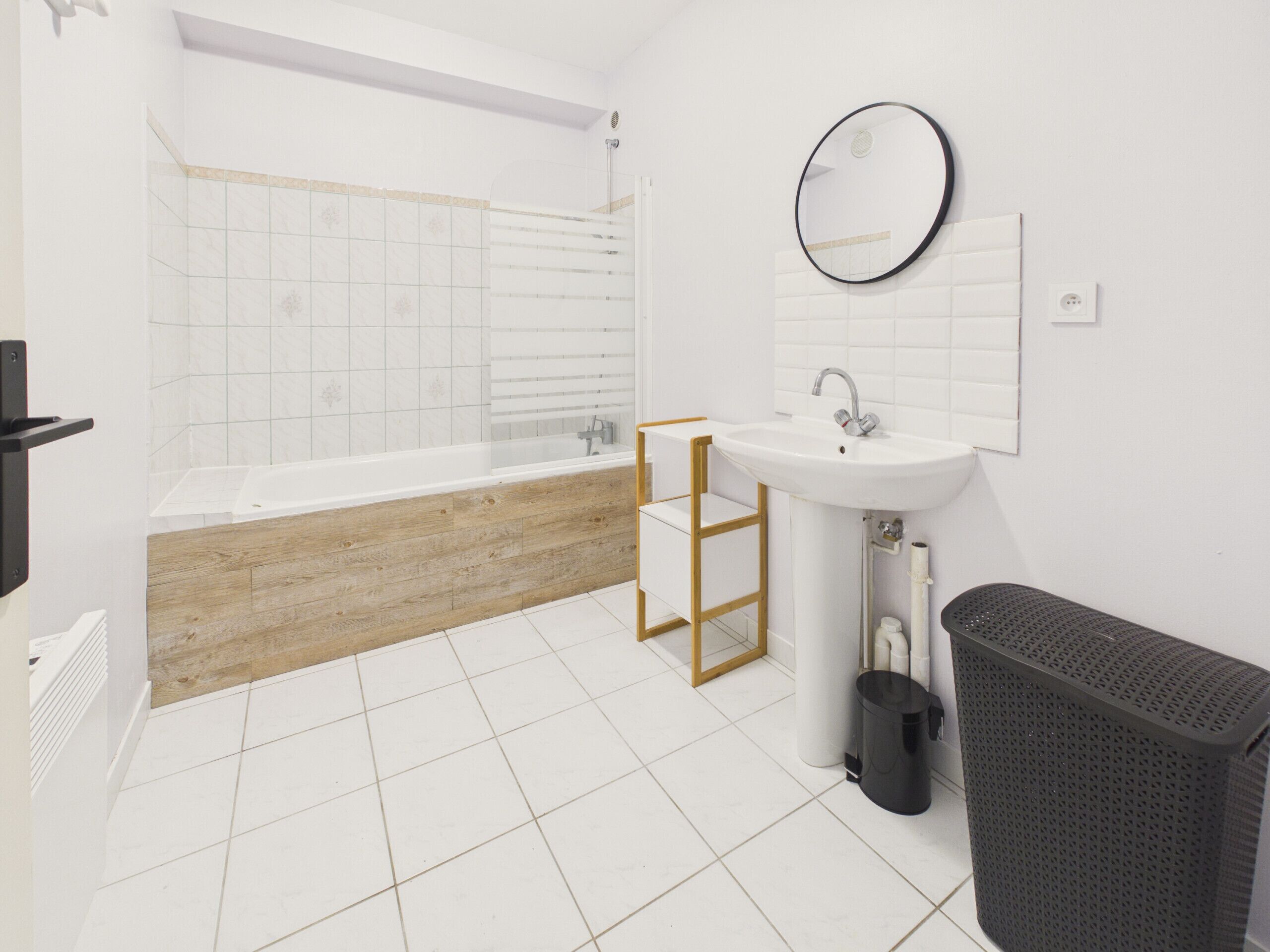 A modern bathroom with a bathtub, white sink, wooden vanity, and a black trash bin.