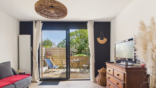 A living room with a sofa, TV, wooden dresser, and a sliding glass door leading to an outdoor patio with a table and chairs. - Near the sea, apartment for 2 with terrace (Le Croisic)