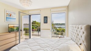 Un dormitorio con una cama grande, un televisor sobre un aparador de madera, y un balcón con vistas a la vegetación y una masa de agua.