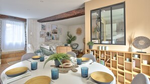Een eetgedeelte met een ronde tafel, blauw glaswerk en houten schalen. Er staan stoelen, een boekenkast en een grote spiegel aan de muur.