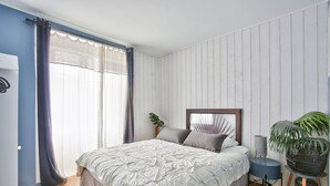 Een slaapkamer met een bed, gordijnen, een plant en een ronde tafel.