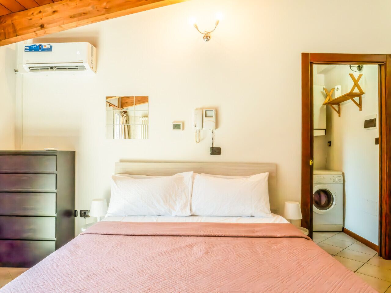 Een slaapkamer met een bed, een dressoir, een airconditioner, een aan de muur bevestigd telefoontoestel, een wasmachine en een houten plafond.