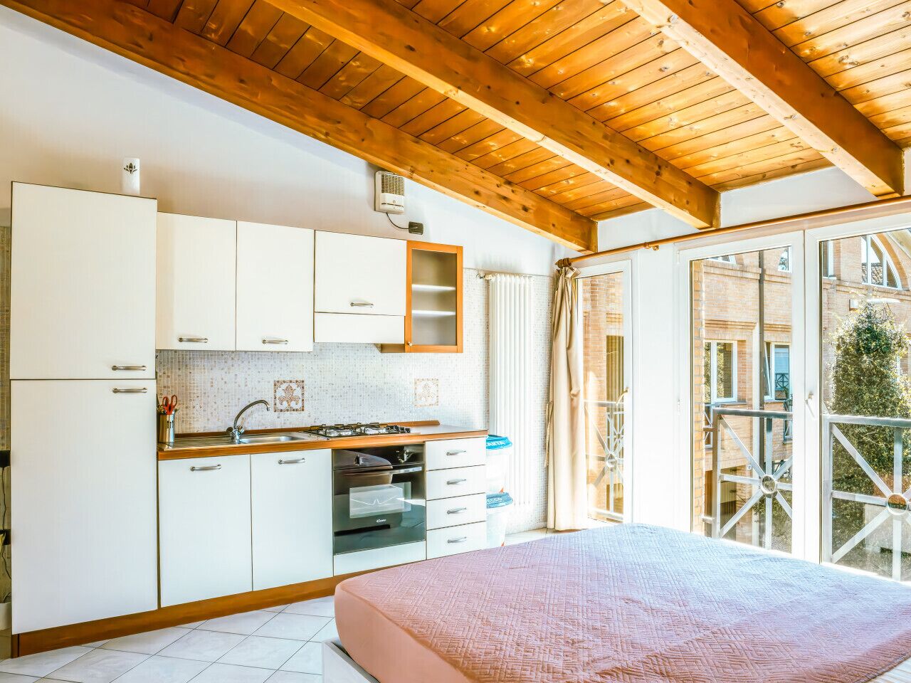 Une chambre avec une grande fenêtre, un plafond en bois et un coin cuisine doté de meubles blancs et d’une cuisinière.