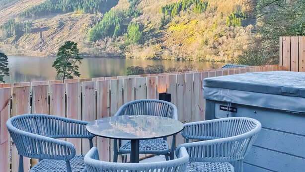 Una terrazza con sedie in vimini e un tavolo che si affaccia su un lago e dolci colline boscose.