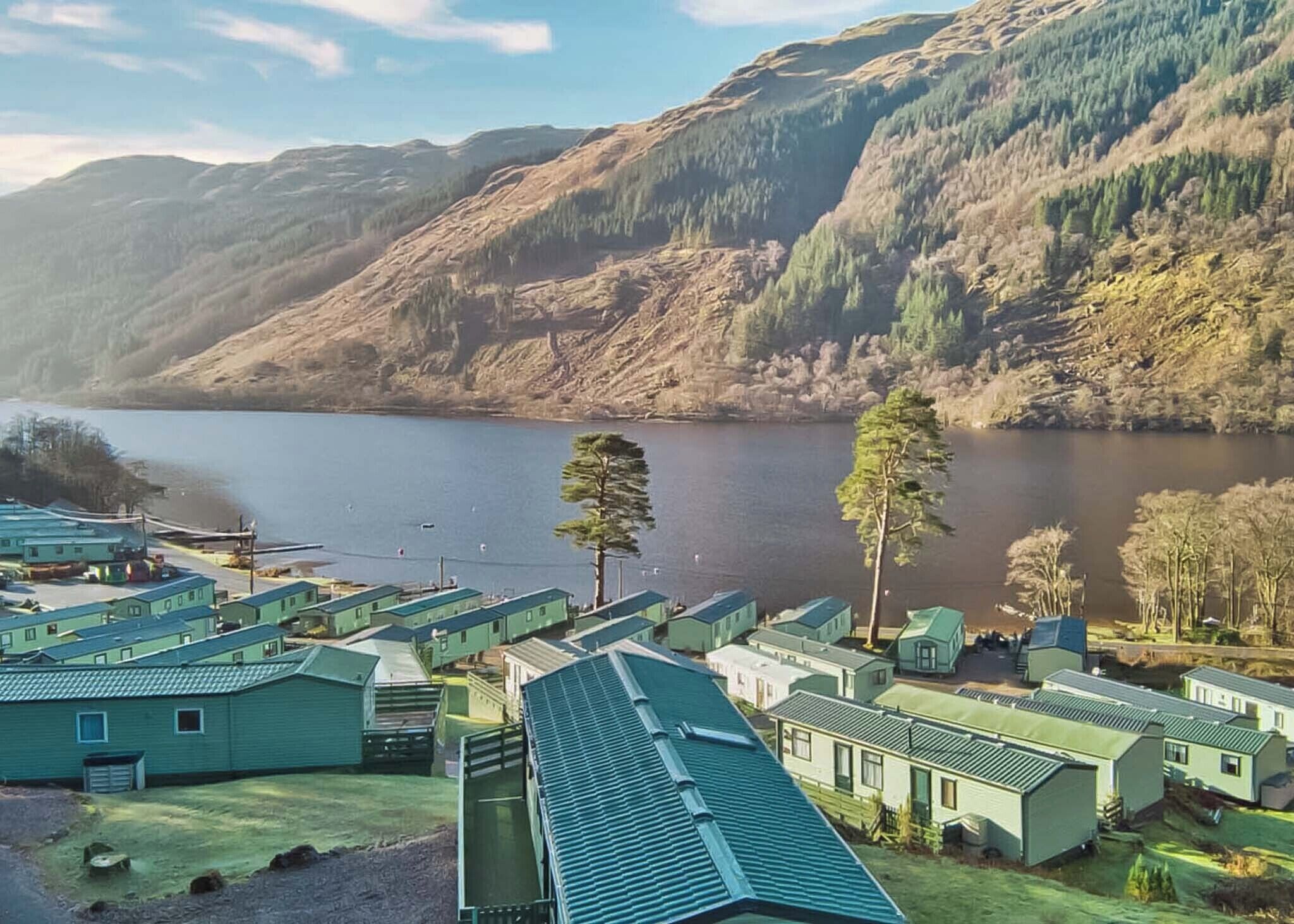 A lakeside view of a campground with multiple green-roofed cabins, a clear sky, and surrounding hills.