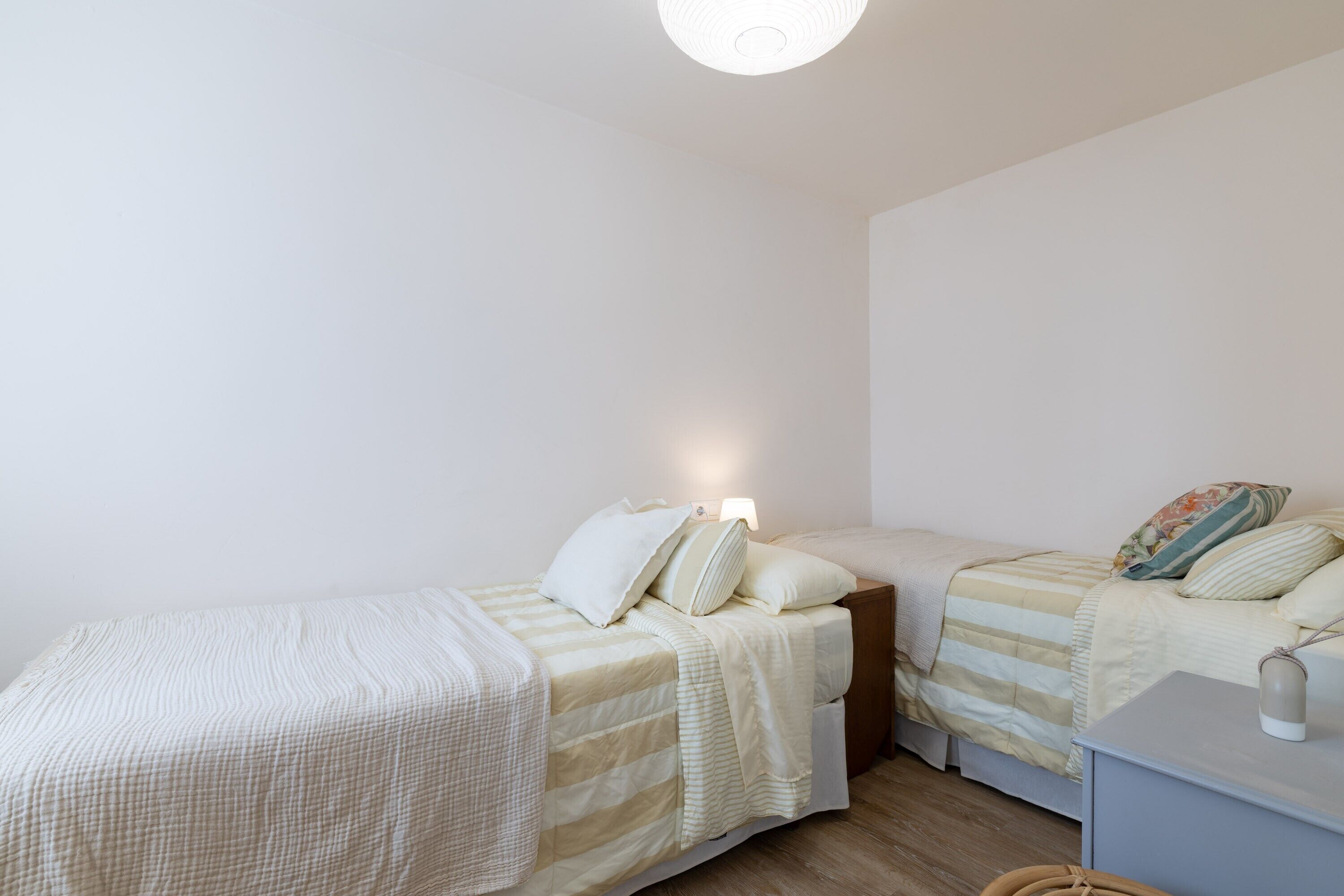 Quarto com duas camas, uma mesinha de cabeceira, um abajur e uma pequena escrivaninha com luminária.