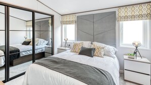 A modern bedroom with a large bed, a mirror, bedside tables, and a window with patterned curtains.