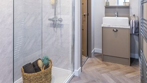 Une salle de bain avec une douche à parois de verre, un panier en bois contenant des articles de toilette, un lavabo blanc et un sol carrelé en motif chevrons.
