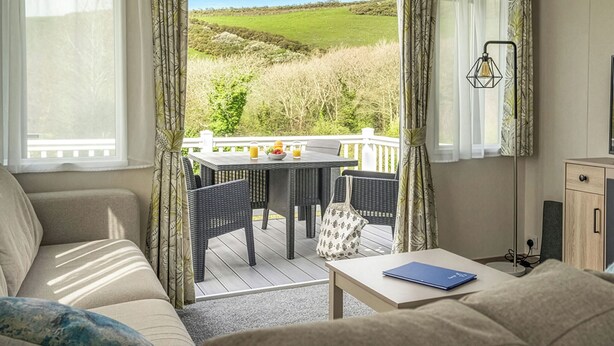 A room with a sofa, a table with chairs, a view of a green landscape, and a balcony with a railing. - 1 bedroom accommodation in Mawgan Porth, Nr Newquay (Mawgan Porth, Nr Newquay)