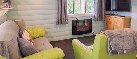 A cozy living room with a sofa, a green armchair, a flat-screen TV, a fireplace, and a window with curtains.