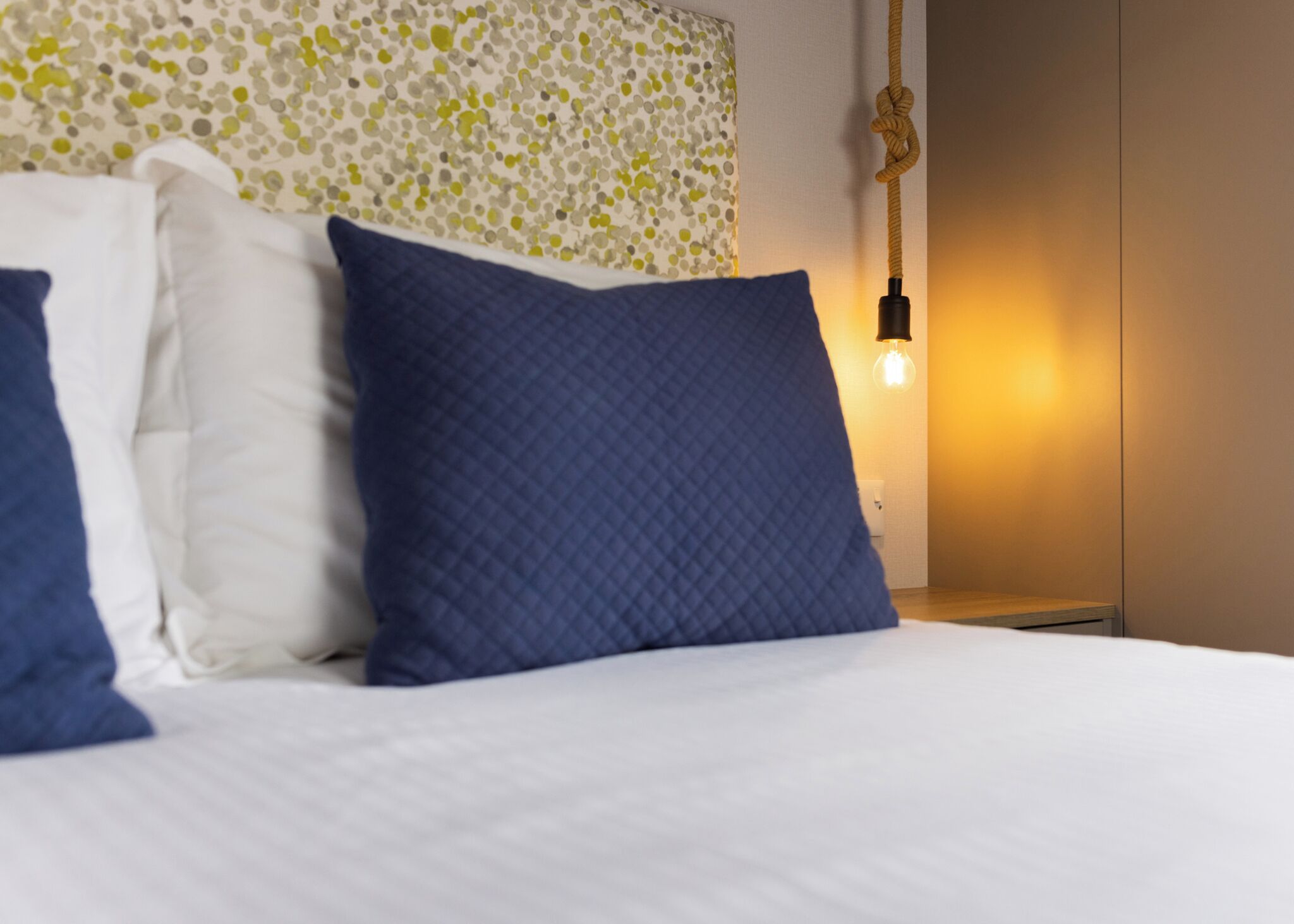 A neatly made bed with white and blue pillows, a textured wall headboard, and a hanging light fixture.