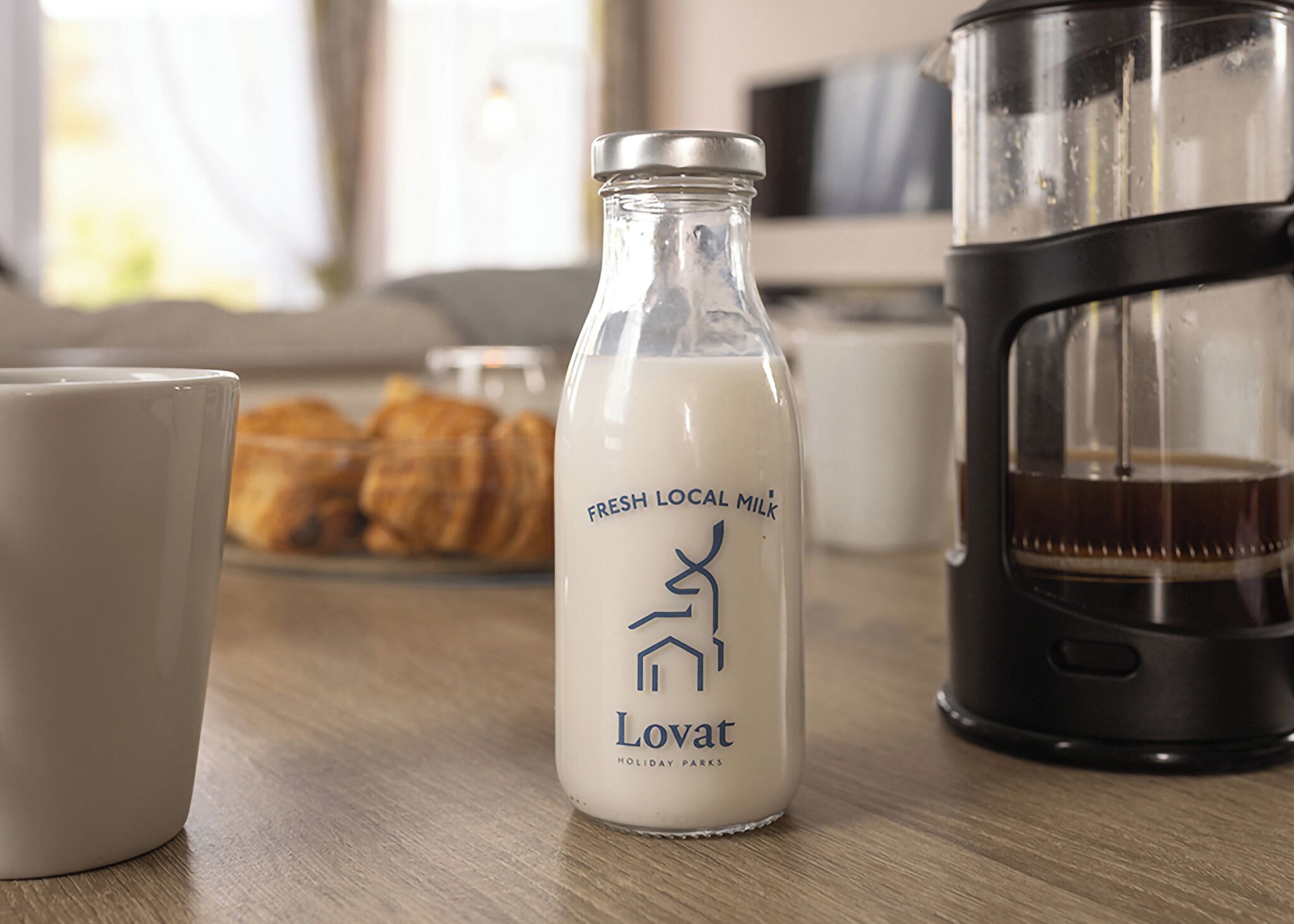 Una bottiglia di latte fresco locale, una macchina da caffè e un piatto di croissant su un tavolo di legno.