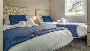 Ein Hotelzimmer mit zwei Betten, jeweils mit blau-weißer Bettwäsche, einem Nachttisch mit Lampe und einem Fenster mit Blick auf ein Haus.