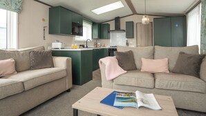 A compact living space with a kitchenette, two sofas, a coffee table with magazines, and a window with curtains. - 1 bedroom accommodation in Mawgan Porth, Nr Newquay (Mawgan Porth, Nr Newquay)