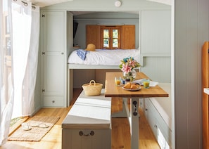 A compact room with a bunk bed, a small table with breakfast items, a wicker basket, and a bench. - Coastal Hideaway Shepherds Hut (Mawgan Porth, Nr Newquay)