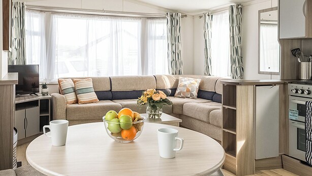 A modern living room with a sofa, a dining table with fruit and coffee cups, a kitchen area with appliances, and large windows with curtains. - Standard 2 Bed (Pet) (Mawgan Porth, Nr Newquay)
