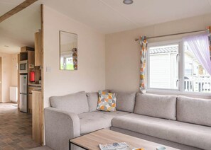 A living room with a sofa, a coffee table, a mirror, and a window with curtains. - The Superior 2 Bedroom is a comfortable and spacious sleeps 6 caravan. (Mawgan Porth, Nr Newquay)