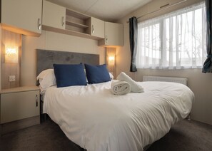 A hotel room with a large bed, two bedside tables, a headboard, and a window with curtains. - The Superior 2 Bedroom is a comfortable and spacious sleeps 6 caravan. (Mawgan Porth, Nr Newquay)