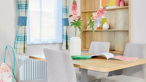 Een eetgedeelte met een houten tafel, vier grijze stoelen, een vaas met roze bloemen, een boek en een roze tas.