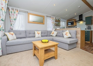A modern living room with a grey sectional sofa, a wooden coffee table, and a kitchen area with wooden cabinets and a TV. - 3 bedroom accommodation in Dawlish (Dawlish)