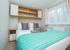 A bedroom with a bed, a bedside table, a nightstand, a built-in shelf with flowers, a window with curtains, and a radiator. - 3 bedroom accommodation in Dawlish (Dawlish)