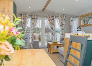 A cozy room with a wooden table, a grey sofa, and a small table with a bowl. There are curtains with a floral pattern, a balcony with a railing, and a window with a view of the outside. - 3 bedroom accommodation in Dawlish (Dawlish)