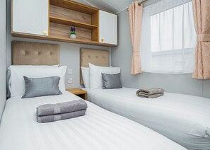 A bedroom with two beds, a wooden shelf with a potted plant, a window with curtains, and a bedside table with a lamp. - 3 bedroom accommodation in Dawlish (Dawlish)