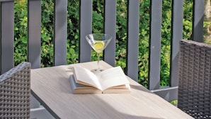 A wooden table with an open book and a martini glass with a lemon slice on a balcony.