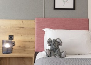 A neatly made bed with a white pillow and a teddy bear, a wooden nightstand with a lamp, and a framed picture on the wall.