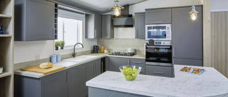 A modern kitchen with grey cabinets, a marble countertop, a built-in oven, and a central island with a bowl of apples.