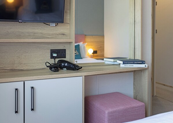 Une chambre d’hôtel avec un bureau en bois, un sèche-cheveux noir, une télévision et un miroir.