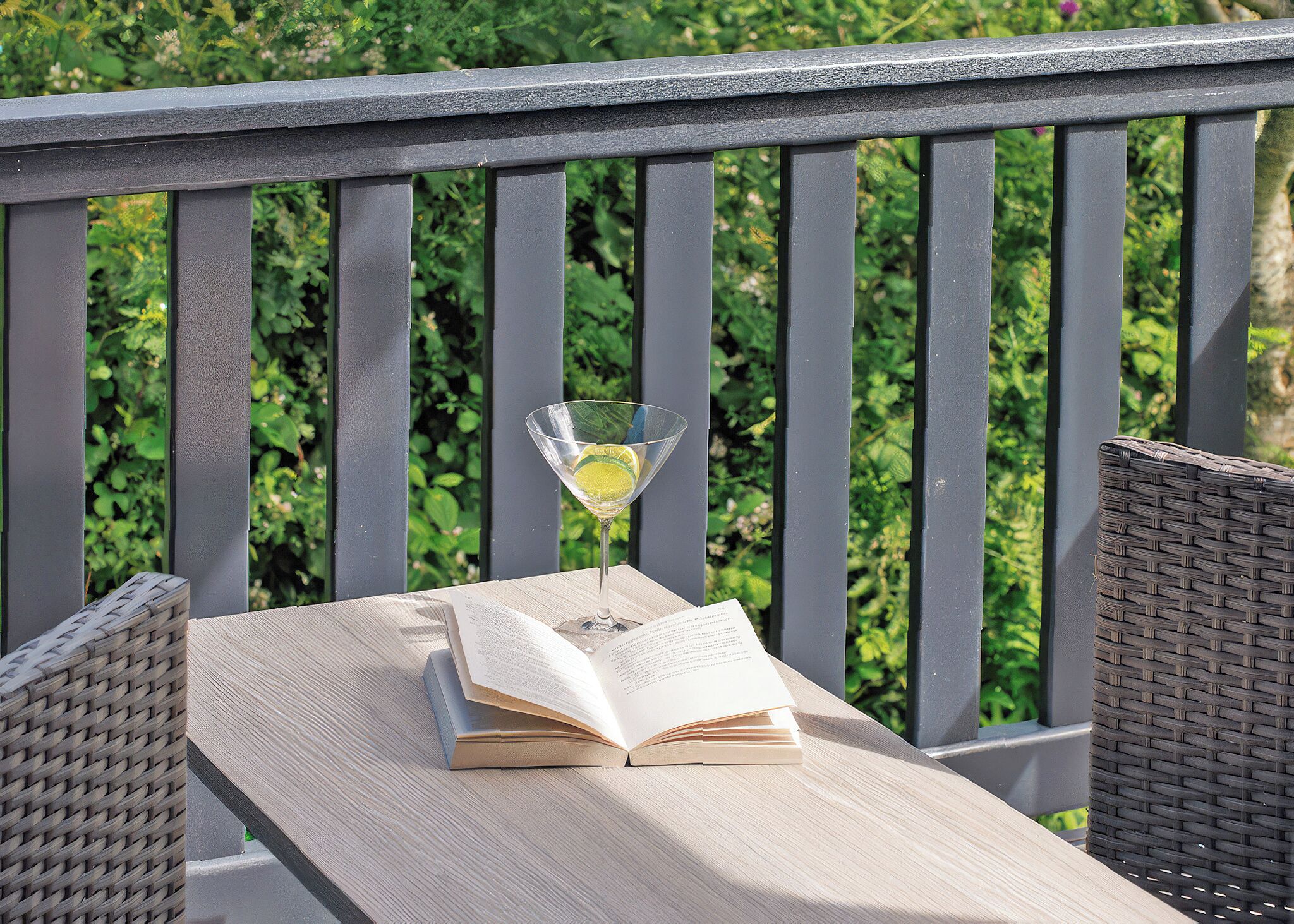 A wooden table with an open book and a martini glass with a lemon slice on a balcony.