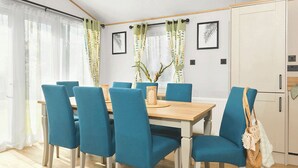 Un coin repas avec une table en bois et des chaises bleues, une fenêtre avec des rideaux et un espace cuisine avec des placards.
