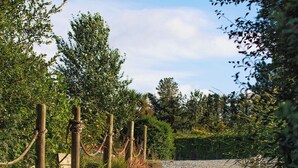 Un chemin de gravier traverse un jardin agrémenté de poteaux et de cordes en bois, le tout étant entouré de verdure et d’arbres.
