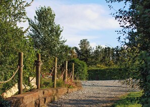 Ein Kiesweg führt durch einen Garten mit Holzpflöcken und Seilen, umgeben von Grünpflanzen und Bäumen.