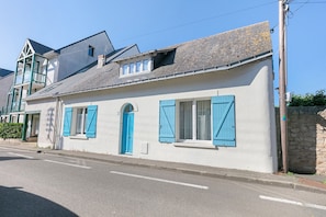 Un bâtiment à deux étages, doté d’une façade blanche, de volets bleus et d’un mur en pierre qui jouxte la propriété.