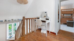 Et værelse med skrivebord i træ, en hvid stol, en fletlampe, et vindue med udsigt og en køjeseng i træ.