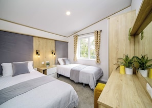A modern bedroom with two beds, a window with curtains, bedside tables, and a small plant. - Exclusive Lodge 3 Bed (Pet) (Padstow)