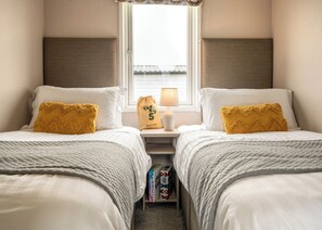 A room with two beds, a bedside table with a lamp, a window, and a bookshelf with books. - 2 bedroom accommodation in Padstow (Padstow)