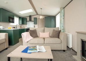 A modern living room with a sofa, a coffee table with magazines, a kitchen area with green cabinets, and a fireplace. - 1 bedroom accommodation in Padstow (Padstow)