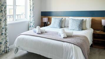 A neatly made bed with white linens and a knitted blanket, a wooden headboard, and a window with floral curtains.