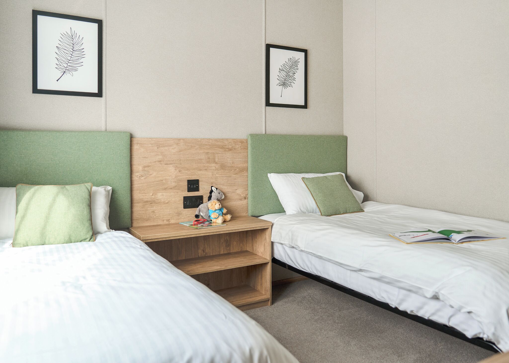Une chambre d’hôtel avec deux lits, des têtes de lit vertes, du linge de lit blanc, une table de chevet avec un ours en peluche, et des œuvres d’art représentant des feuilles, encadrées et accrochées au mur.